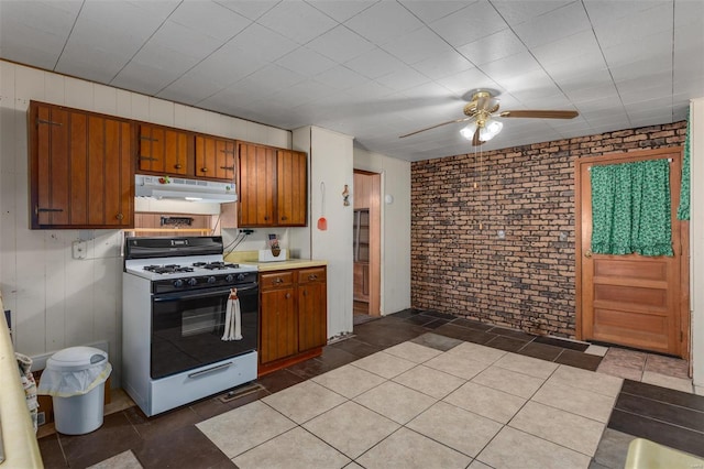 kitchen featuring brown cabinets, under cabinet range hood, range with gas stovetop, brick wall, and ceiling fan