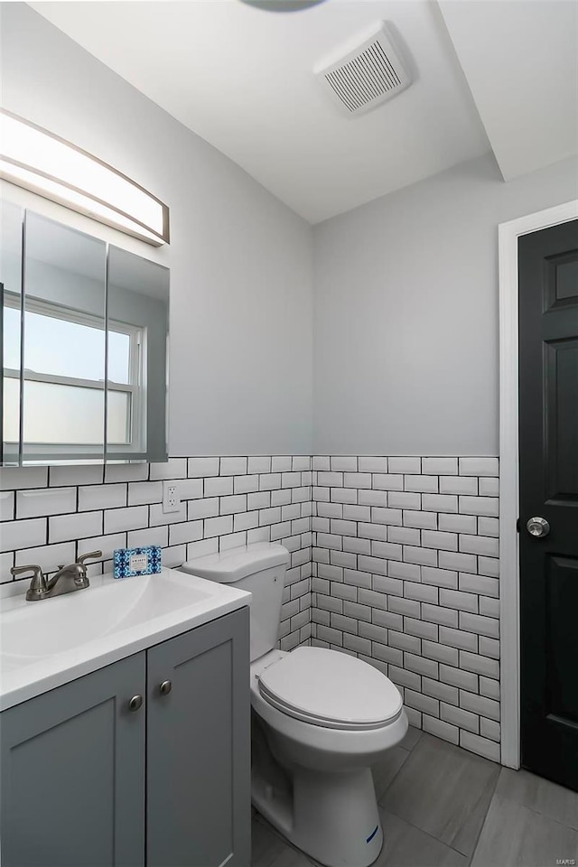 half bathroom featuring visible vents, tile walls, a wainscoted wall, toilet, and vanity