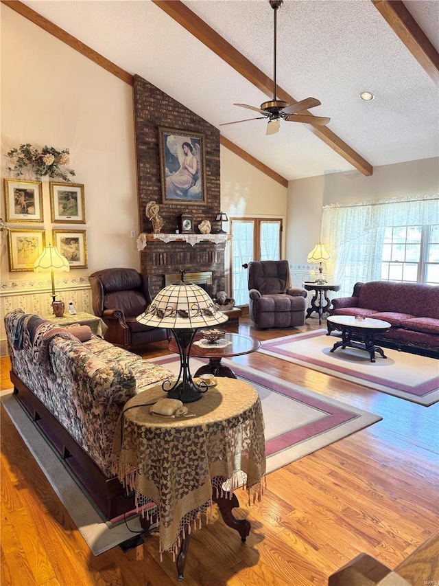 living room with a brick fireplace, ceiling fan, lofted ceiling with beams, wood finished floors, and a textured ceiling