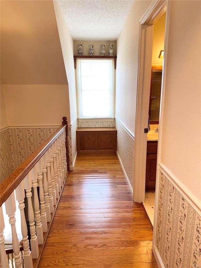 hall with light wood finished floors, a textured ceiling, wainscoting, and wallpapered walls