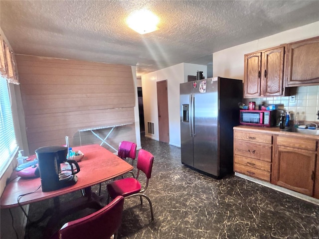 kitchen with stainless steel refrigerator with ice dispenser, a sink, backsplash, wood walls, and brown cabinetry