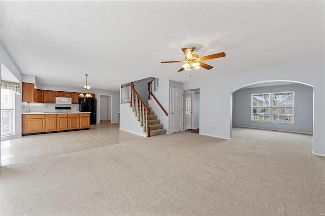 unfurnished living room featuring a ceiling fan, arched walkways, stairs, a textured ceiling, and light colored carpet