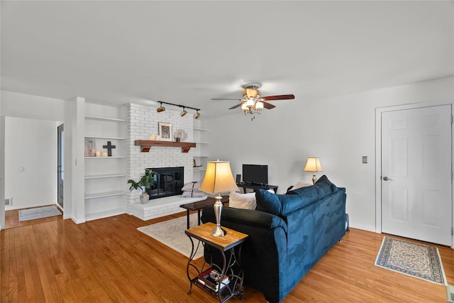 living room with track lighting, a brick fireplace, baseboards, ceiling fan, and light wood-type flooring