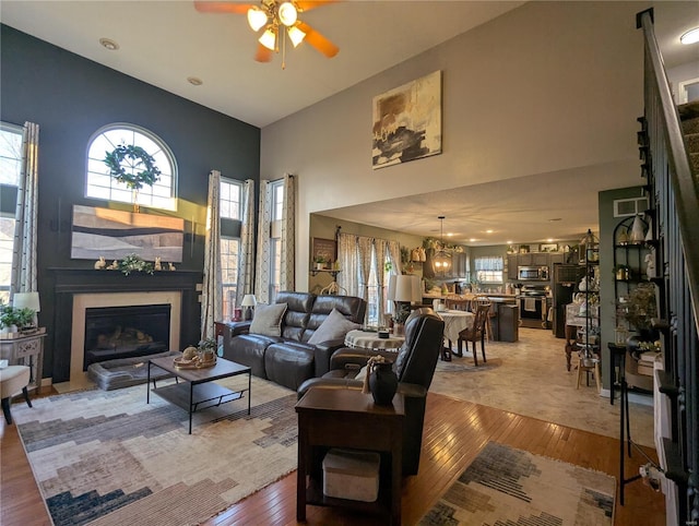 living area with a glass covered fireplace, ceiling fan with notable chandelier, light wood-style floors, and a towering ceiling