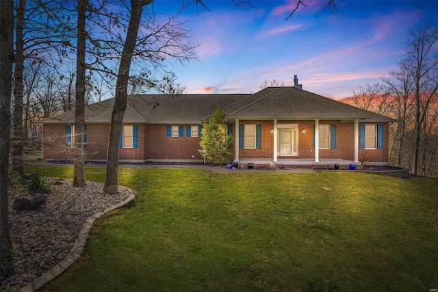 back of house at dusk featuring a yard, brick siding, covered porch, and a shingled roof