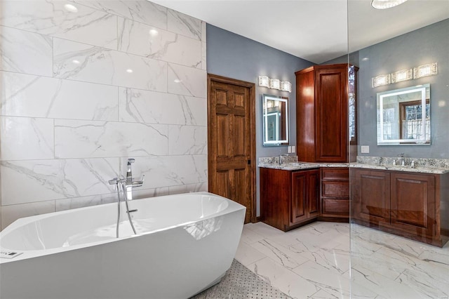 full bath with a freestanding bath, double vanity, marble finish floor, tile walls, and a sink