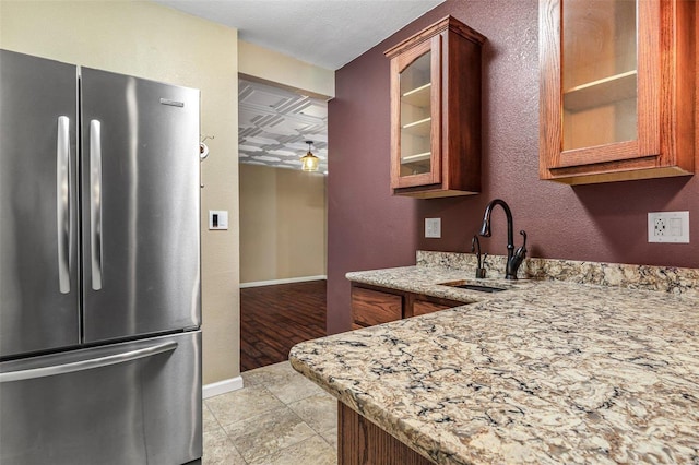 kitchen with light stone countertops, freestanding refrigerator, a sink, glass insert cabinets, and brown cabinets