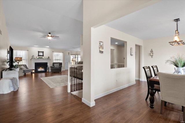 dining area featuring dark wood-type flooring, a ceiling fan, baseboards, and a warm lit fireplace