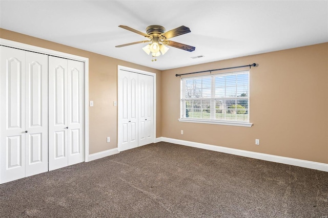 unfurnished bedroom with a ceiling fan, visible vents, baseboards, dark colored carpet, and two closets