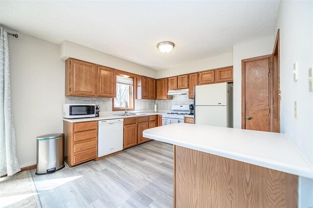 kitchen with white appliances, a peninsula, a sink, under cabinet range hood, and backsplash