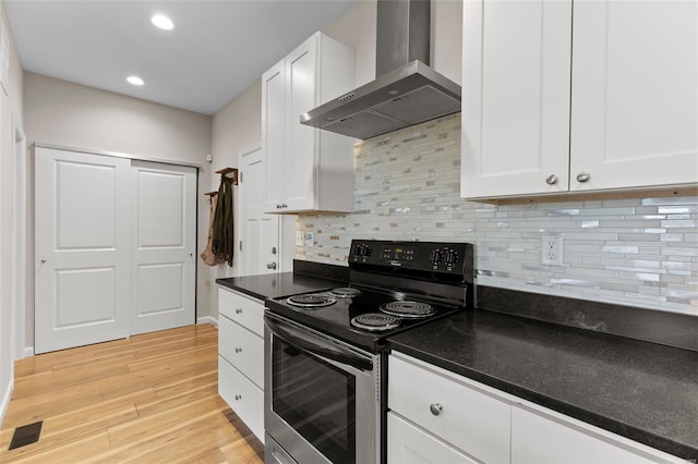 kitchen featuring wall chimney range hood, stainless steel electric range oven, white cabinetry, visible vents, and light wood-style floors