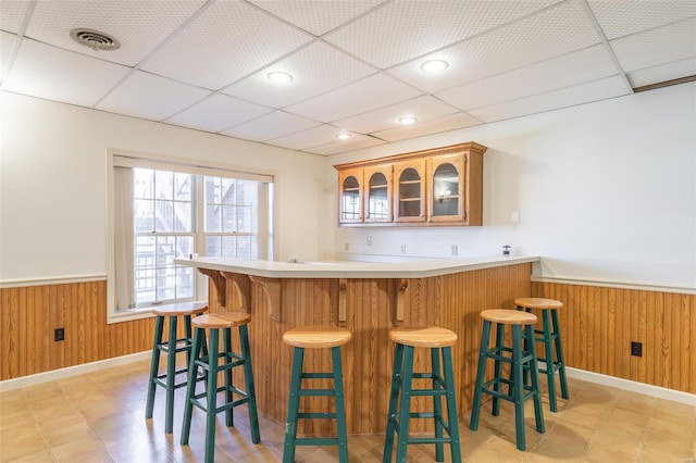 bar featuring wood walls, light floors, wainscoting, visible vents, and a drop ceiling