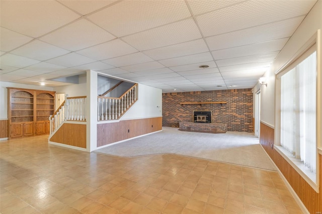 unfurnished living room with wood walls, stairway, wainscoting, a fireplace, and a paneled ceiling