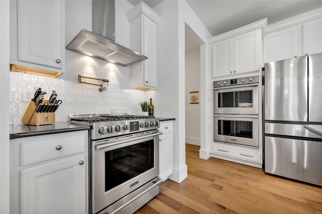 kitchen with wall chimney exhaust hood, dark countertops, appliances with stainless steel finishes, light wood-style flooring, and decorative backsplash