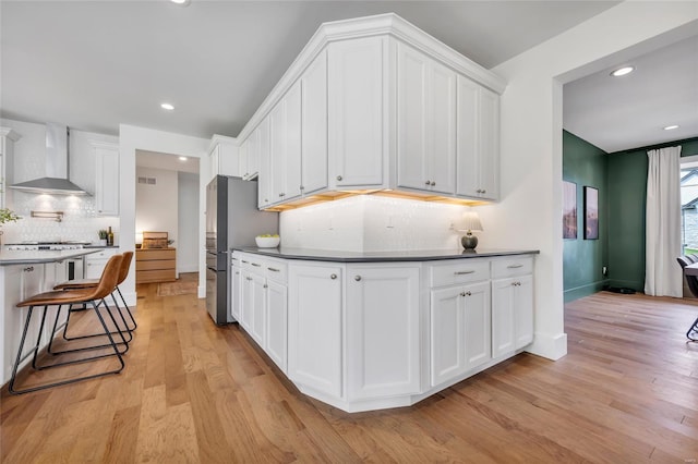 kitchen featuring freestanding refrigerator, light wood-type flooring, wall chimney range hood, white cabinetry, and recessed lighting