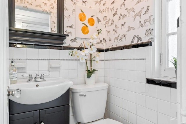 bathroom with tile walls, wainscoting, vanity, toilet, and wallpapered walls