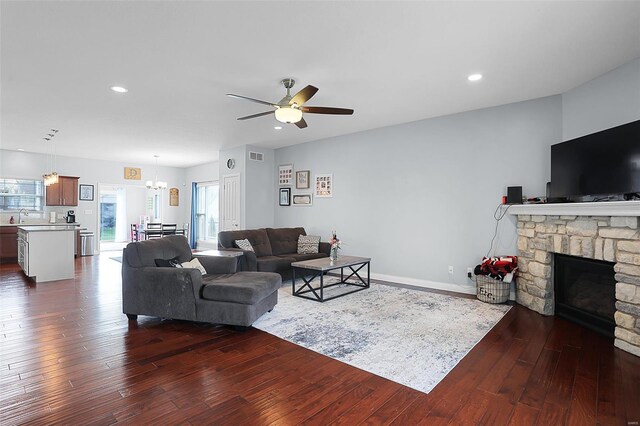 living room with dark wood-type flooring, a stone fireplace, ceiling fan with notable chandelier, visible vents, and recessed lighting