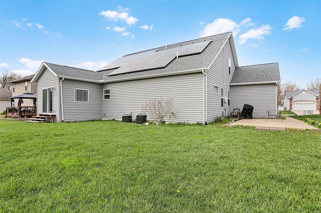 back of property with roof mounted solar panels, a patio, a deck, roof with shingles, and a lawn