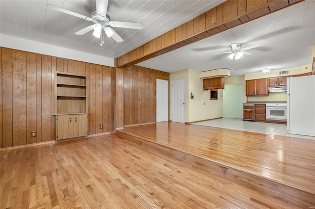 unfurnished living room with light wood-style floors, a ceiling fan, visible vents, wooden walls, and baseboards