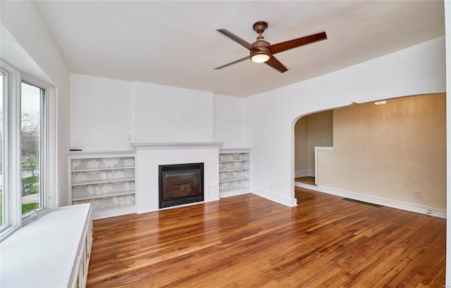unfurnished living room featuring ceiling fan, a glass covered fireplace, wood finished floors, and a healthy amount of sunlight