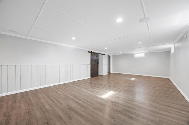 basement featuring baseboards, crown molding, a wainscoted wall, a barn door, and wood finished floors