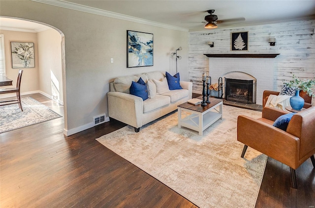 living area featuring arched walkways, a brick fireplace, a ceiling fan, crown molding, and wood finished floors