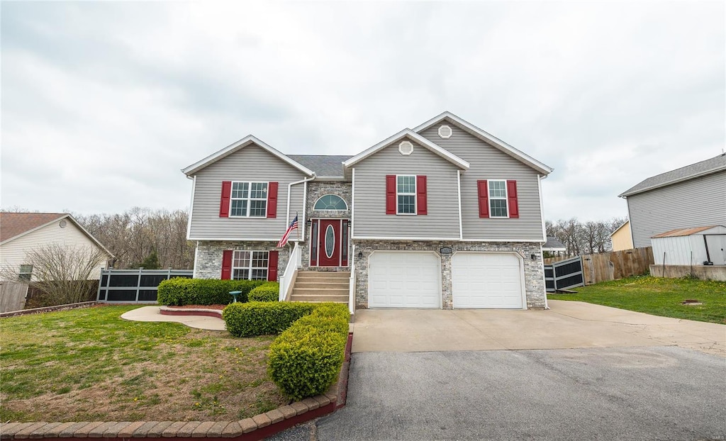raised ranch featuring concrete driveway, stone siding, a front lawn, an attached garage, and fence