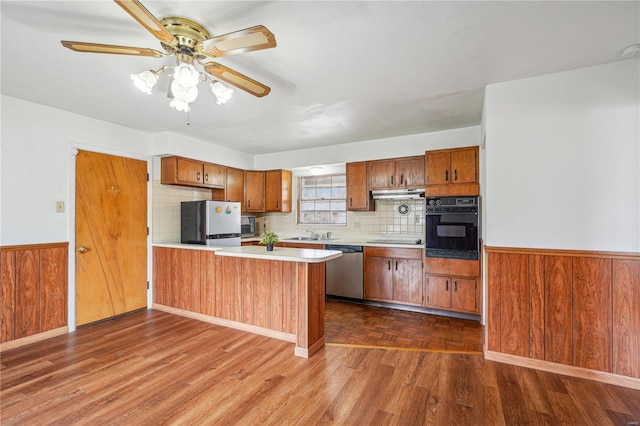 kitchen featuring light countertops, appliances with stainless steel finishes, a peninsula, brown cabinets, and wainscoting