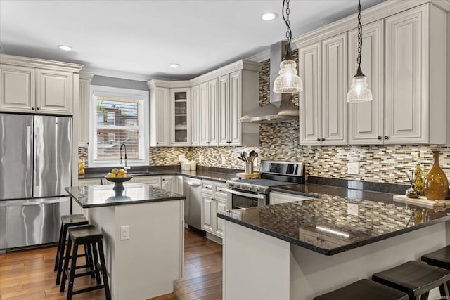 kitchen featuring wall chimney exhaust hood, stainless steel appliances, a peninsula, a sink, and a kitchen bar