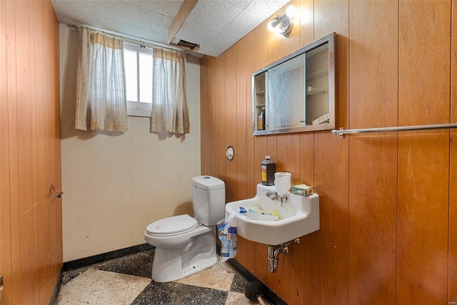 half bathroom featuring wood walls, toilet, a sink, baseboards, and a paneled ceiling