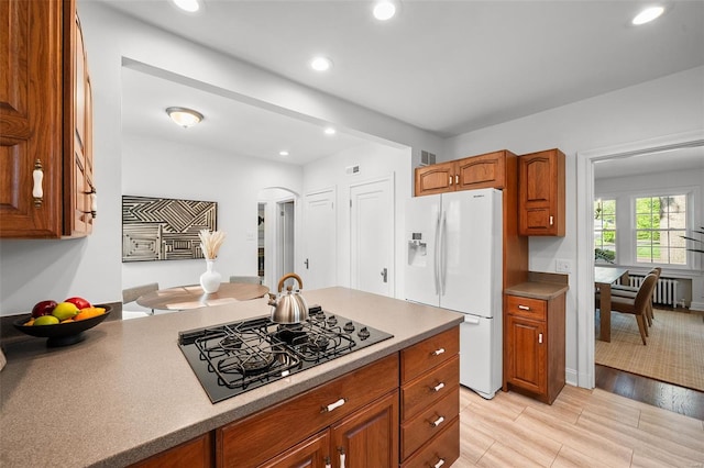 kitchen with brown cabinetry, radiator heating unit, white refrigerator with ice dispenser, black stovetop, and arched walkways