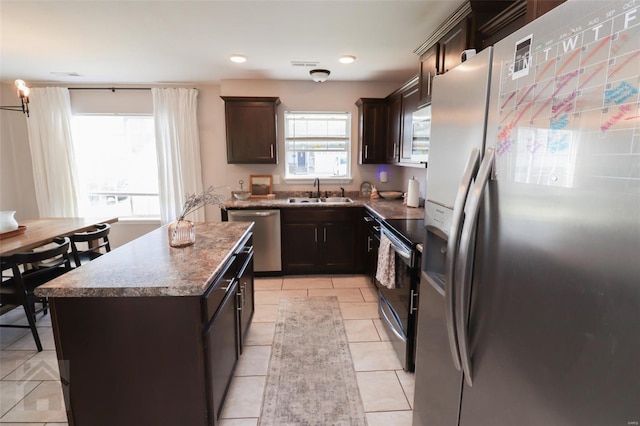 kitchen with stainless steel appliances, dark brown cabinets, a sink, a center island, and dark stone counters