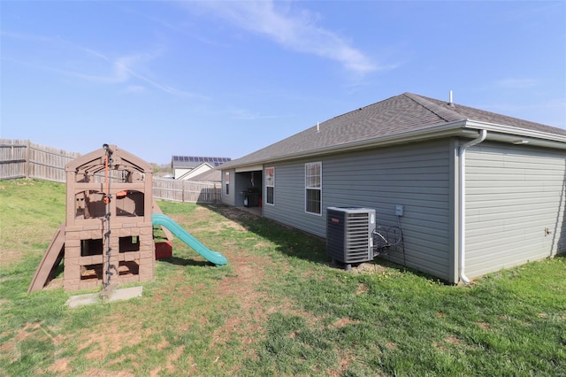 back of house with a playground, a fenced backyard, central AC, a lawn, and roof with shingles