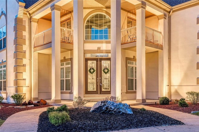 view of exterior entry featuring roof with shingles, covered porch, stucco siding, and a balcony