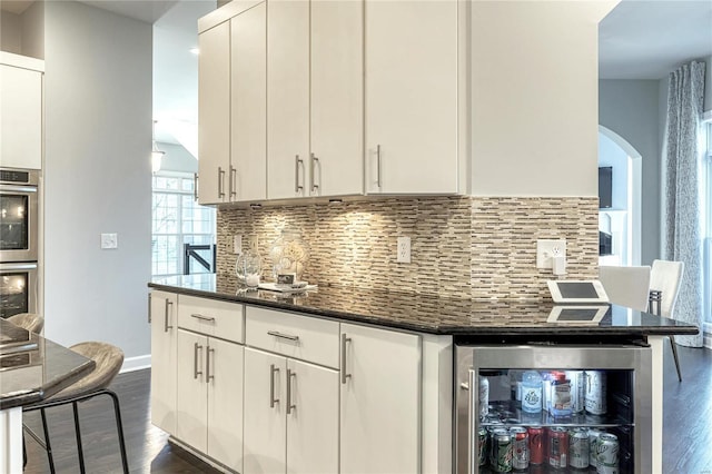 kitchen featuring dark wood-style flooring, stainless steel double oven, beverage cooler, arched walkways, and tasteful backsplash