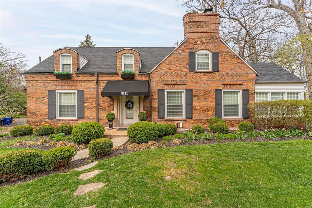view of front of property with a shingled roof, a chimney, brick siding, and a front yard