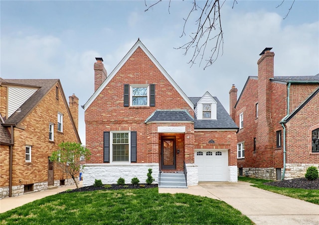 tudor-style house featuring roof with shingles, driveway, a chimney, brick siding, and a front yard