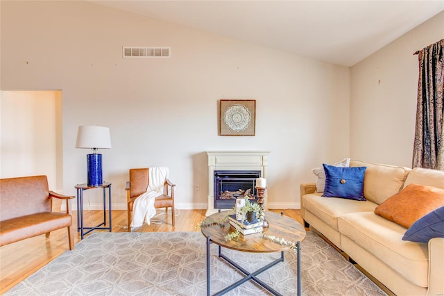 living area featuring wood finished floors, visible vents, vaulted ceiling, baseboards, and a glass covered fireplace