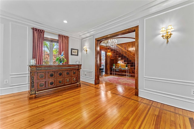 unfurnished room featuring an inviting chandelier, crown molding, stairs, a decorative wall, and light wood-style floors