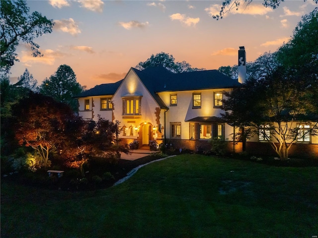 back of house at dusk featuring a lawn, a chimney, and stucco siding