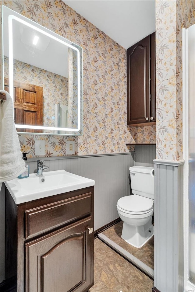 bathroom with wallpapered walls, a wainscoted wall, vanity, and toilet