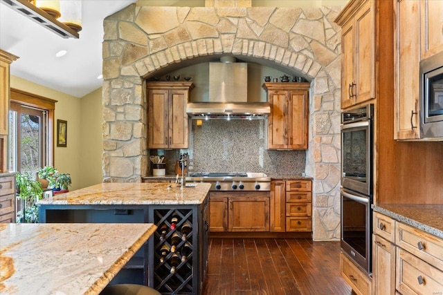kitchen featuring wall chimney range hood, brown cabinets, stainless steel appliances, decorative backsplash, and dark wood-type flooring