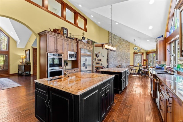 kitchen featuring dark wood-style flooring, a kitchen island with sink, built in appliances, dark cabinetry, and high vaulted ceiling