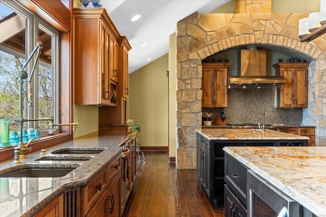 kitchen featuring wall chimney range hood, dark wood-style floors, brown cabinetry, and light stone counters