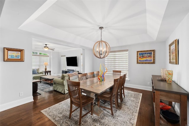 dining space with ceiling fan with notable chandelier, baseboards, a tray ceiling, and dark wood finished floors
