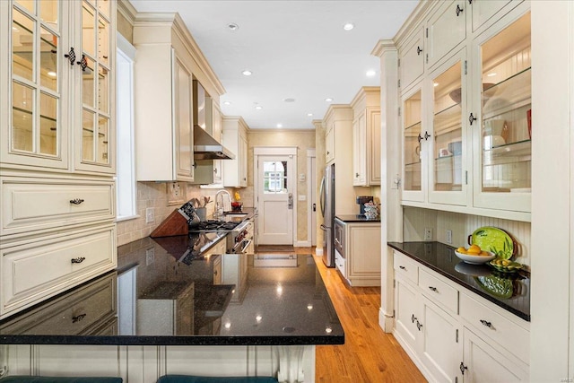 kitchen with appliances with stainless steel finishes, tasteful backsplash, a peninsula, light wood finished floors, and wall chimney exhaust hood