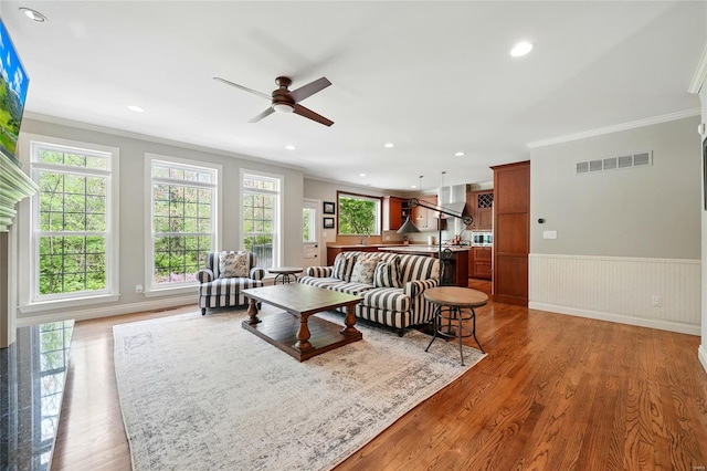 living area with wood finished floors, ceiling fan, a wainscoted wall, ornamental molding, and visible vents