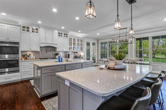 kitchen with dark wood-style flooring, recessed lighting, stainless steel double oven, gray cabinetry, and an island with sink