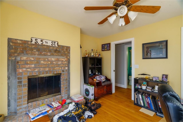 living area featuring baseboards, wood finished floors, ceiling fan, and a brick fireplace