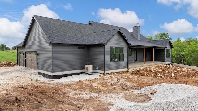 back of house with covered porch, a chimney, a shingled roof, an attached garage, and ac unit
